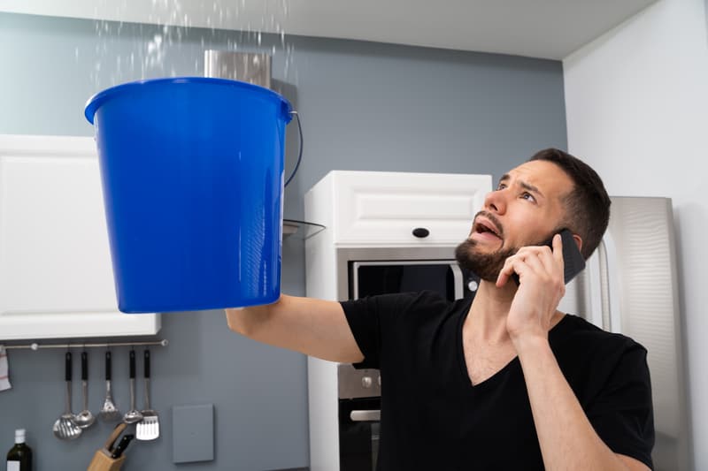 A homeowner on the phone, holding a bucket under a ceiling leak