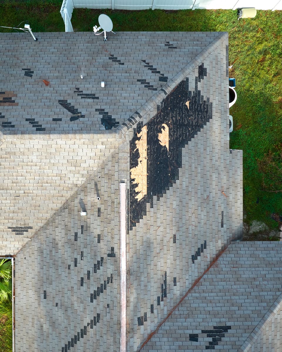 Aerial photo of a storm-damaged roof. One slope is stripped to the black underlayment with bare plywood decking showing, and missing shingle tabs are scattered across the rest.